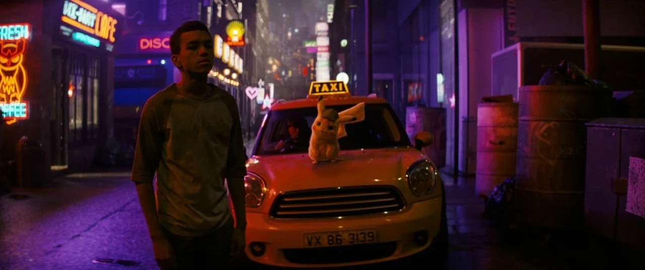 Pokémon Detective Pikachu movie still: fullbody — A neon-lit urban alleyway at night features diverse signage including an owl-themed cafe. A young…