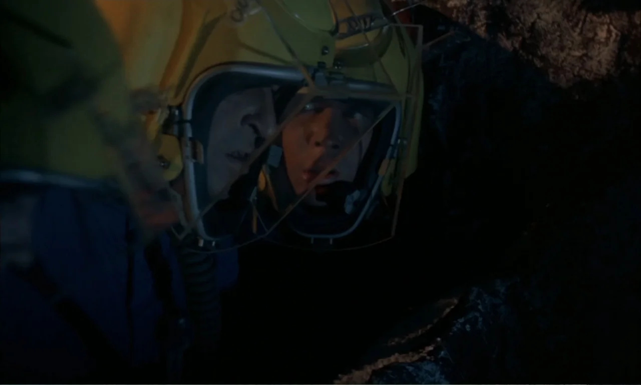 Journey to the Seventh Planet (1962) movie still: closeup — Two individuals wearing yellow flight helmets look closely into a dark, narrow space.…