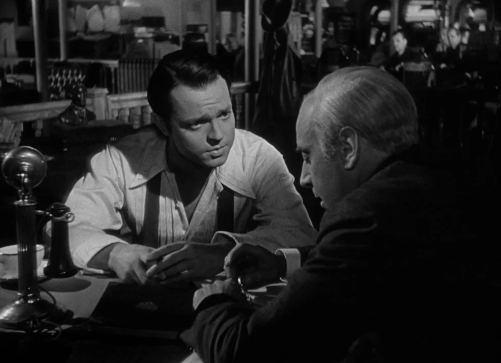 Citizen Kane (1941) movie still: medium — A dimly lit restaurant interior features dark wooden partitions and blurry background silhouettes. Two men…