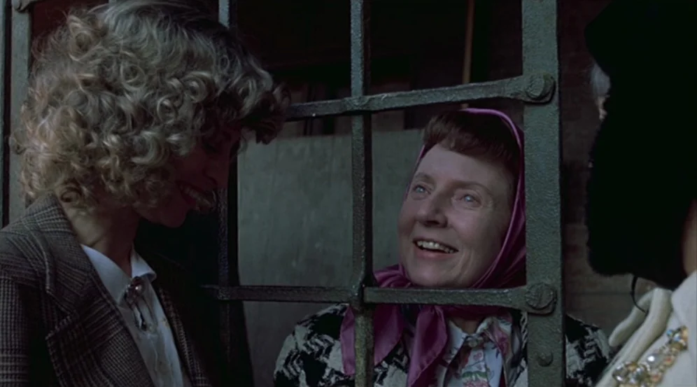 Don’t Look Now (1973) movie still: medium — An older woman in a pink headscarf looks through a black iron gate. A blonde woman standing on the other…