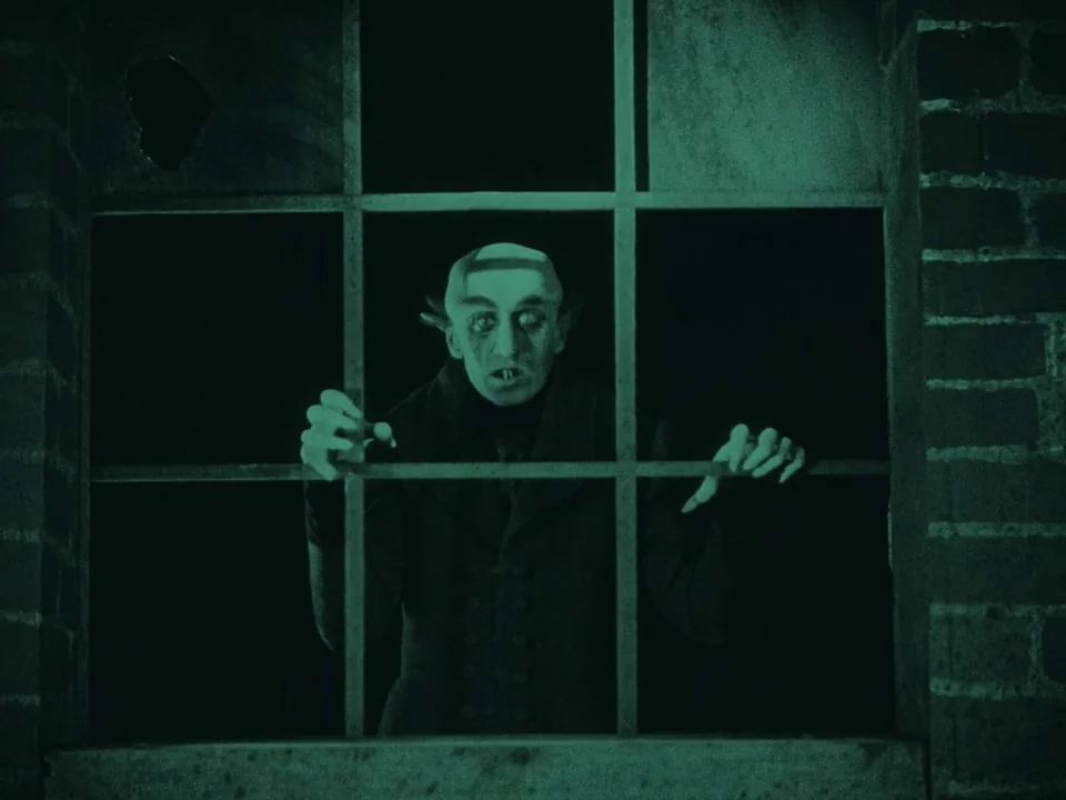 Nosferatu (1922) movie still: medium — A dark, brick-walled building exterior features a gridded pane window at night. A pale, sharp-featured figure…