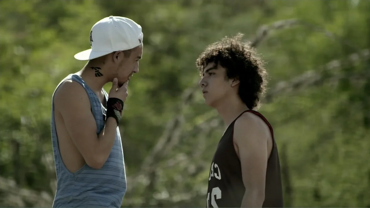 Loco Love (2003) movie still: medium — An outdoor area with blurred green vegetation and trees in the background. Two young men stand facing each…