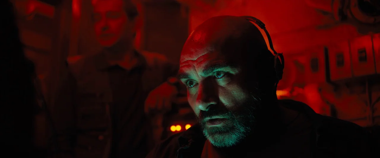 Black Sea (2014) movie still: closeup — A dimly lit industrial environment is bathed in saturated red light and control panels. A bearded man…