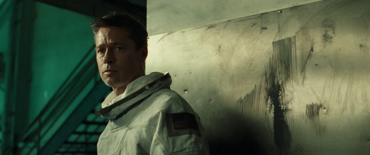 Ad Astra (2019) movie still: medium — A dimly lit industrial hallway features a cold metal wall marked with dark streaks. A man wearing a white…