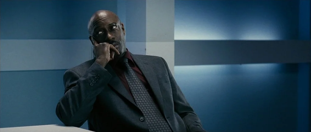 The Walker (2007) movie still: medium — A stark room with blue walls and horizontal white lighting panels along the back. A middle-aged man in a…