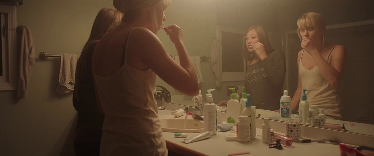 It Follows (2015) movie still: medium — Two women stand in a bathroom at night, illuminated by a warm overhead light. They stand before a large…