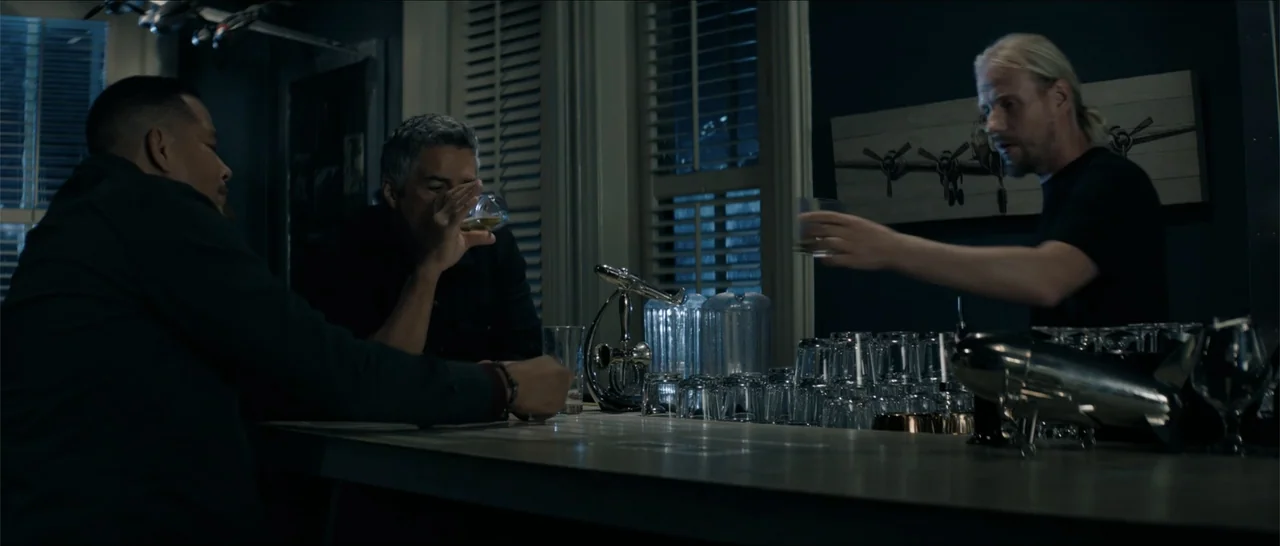Crescent City (2024) movie still: medium — A dimly lit bar interior featuring a wooden counter, glass drinkware, and aviation-themed decor. Three…