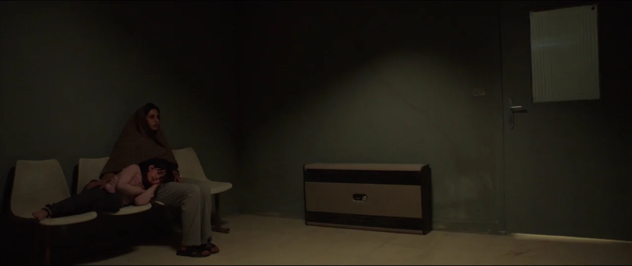Under The Shadow (2016) movie still: wide — A dark, sparsely furnished waiting room has olive-green walls and a single door. An adult woman sits on…