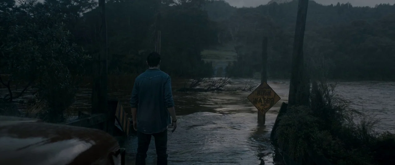 Evil Dead (2013) movie still: fullbody — A rural road is submerged by dark, rising floodwater surrounded by dense forest trees. A man in a blue…