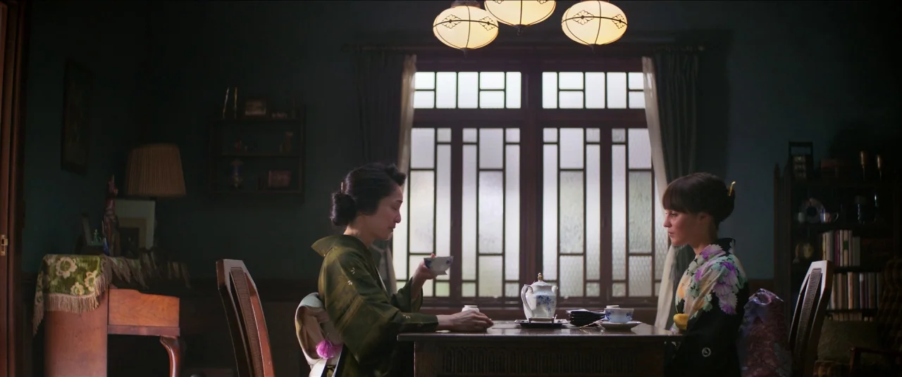 Earthquake Bird movie still: medium — A dimly lit room features a wooden table centered before a large, grid-paneled window. Two women in…