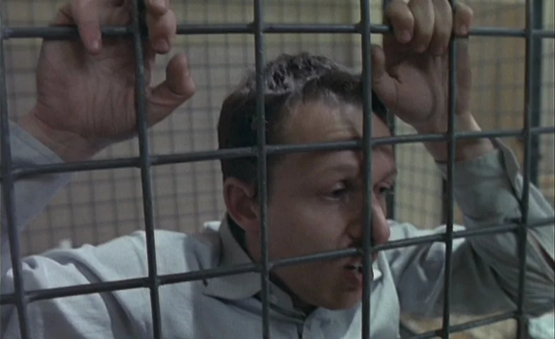 Blind Chance (1987) movie still: closeup — A man stands behind thick iron bars in a brightly lit room. He grips the bars with both hands while…