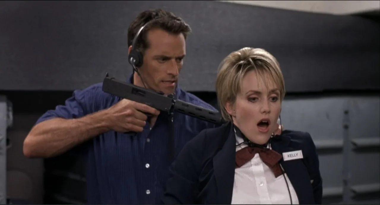 Air Rage (2001) movie still: medium — A dimly lit industrial or office corridor with dark grey walls. A man holds a machine gun at a woman wearing a…