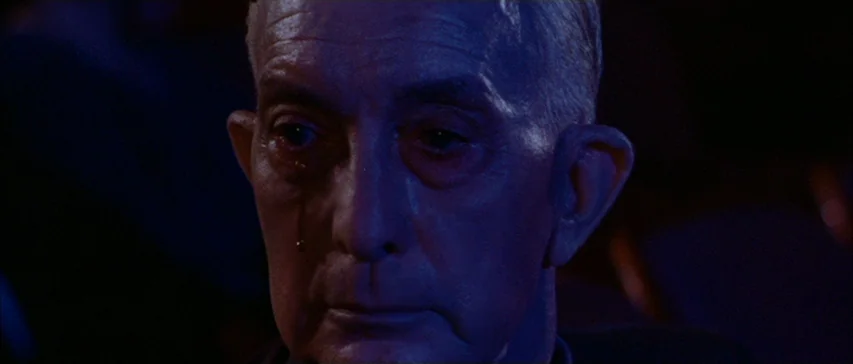 Messiah of Evil (1973) movie still: closeup — An elderly man sits in a dark, dimly lit theater auditorium. A single tear tracks down his cheek while…