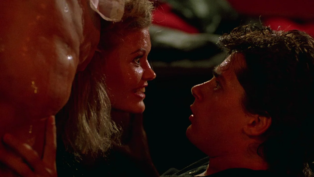 Society (1989) movie still: medium — A dark indoor space provides the setting for a close-up interaction between two people. A woman and a man face…