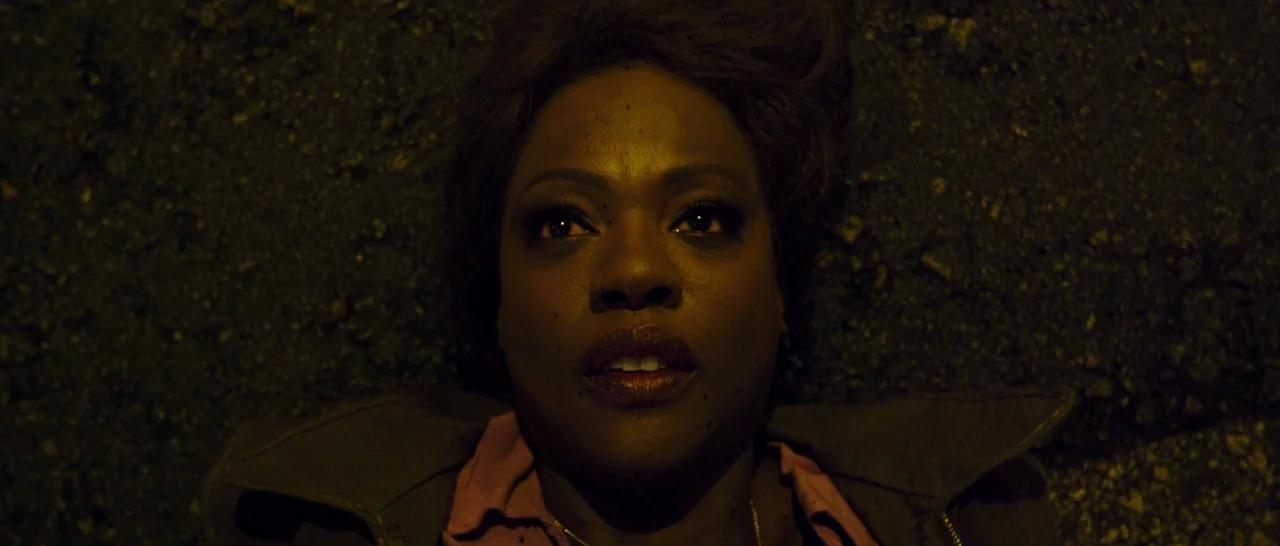 Blackhat (2015) movie still: closeup — A woman lies flat on the dark, grainy surface of an asphalt road at night. She gazes upwards with a vacant,…