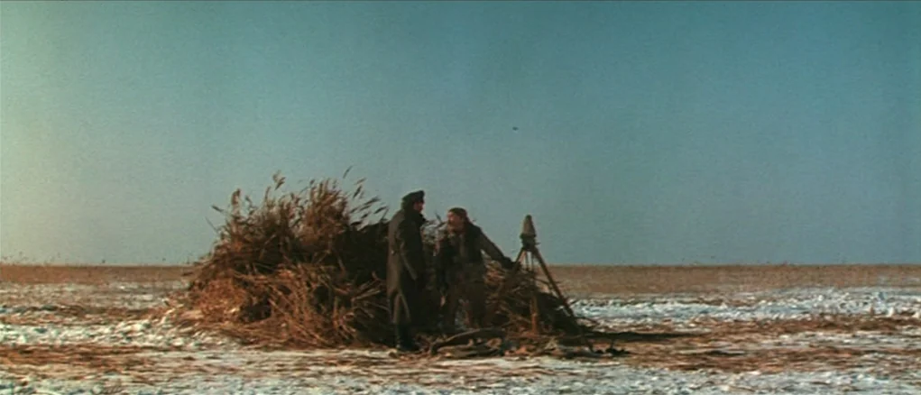 Dersu Uzala (1975) movie still: wide — A flat, snow-covered landscape stretches to the horizon under a clear, pale sky. Two soldiers stand by a pile…