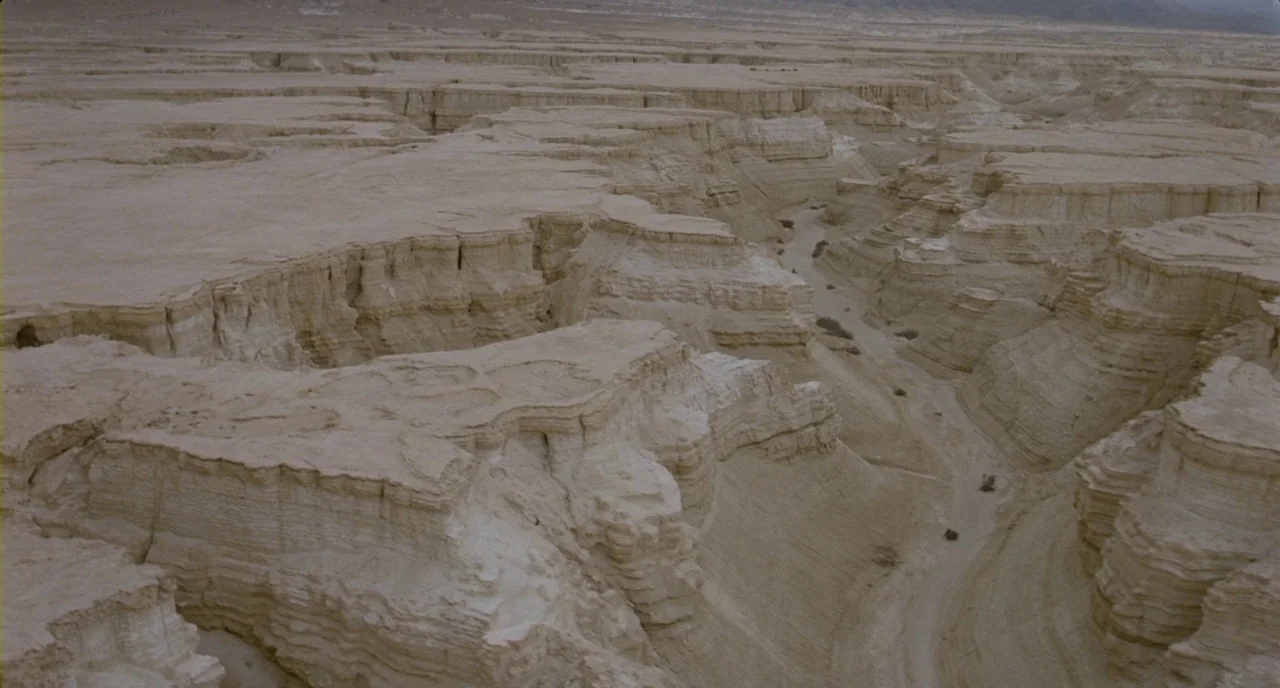 Essential Killing (2010) movie still: establishing — An arid, rocky canyon landscape features layered sedimentary rock formations and dry riverbeds.…
