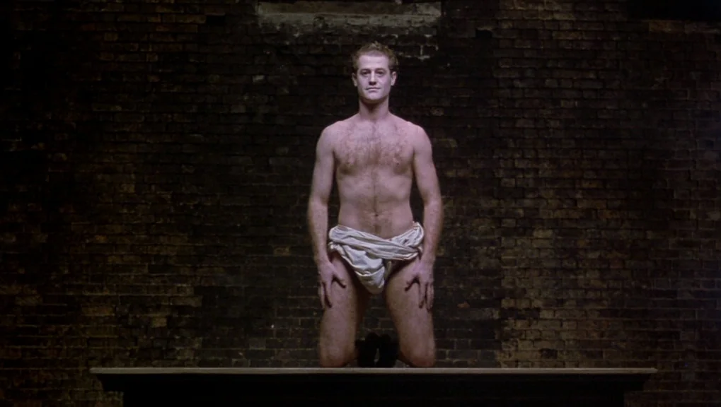 War Requiem (1989) movie still: medium — A man kneels on a dark wooden platform in front of a brick wall. He wears only a draped white cloth around…