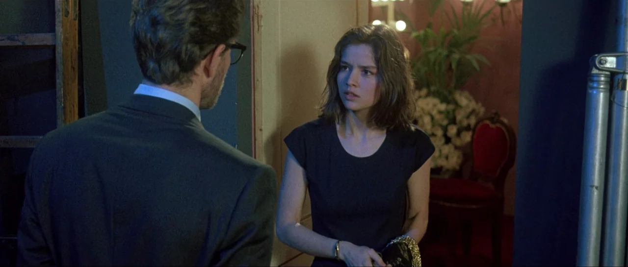 Opera (1987) movie still: medium — A dimly lit room features a red velvet chair and floral arrangements in the background. A woman in a black shirt…