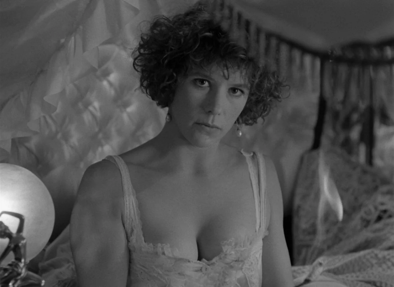 Singapore Sling (1990) movie still: medium — A woman in a lace camisole sits on a bed with a tufted headboard. A bright lamp glows in the foreground…