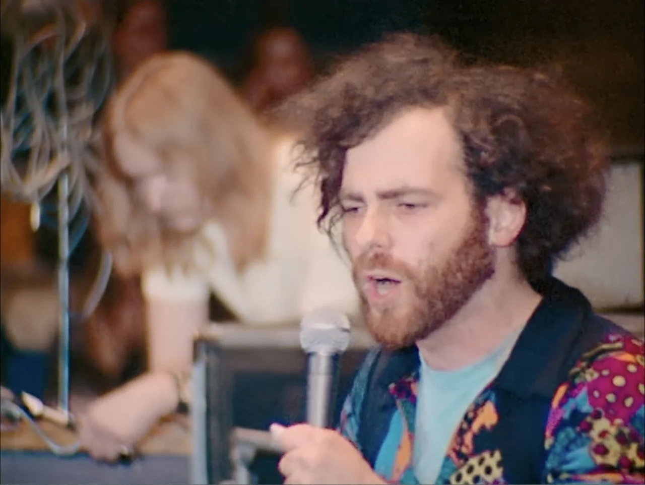 One to One: John & Yoko (2025) movie still: closeup — A dimly lit indoor stage area with music equipment visible in the background. A man with curly…
