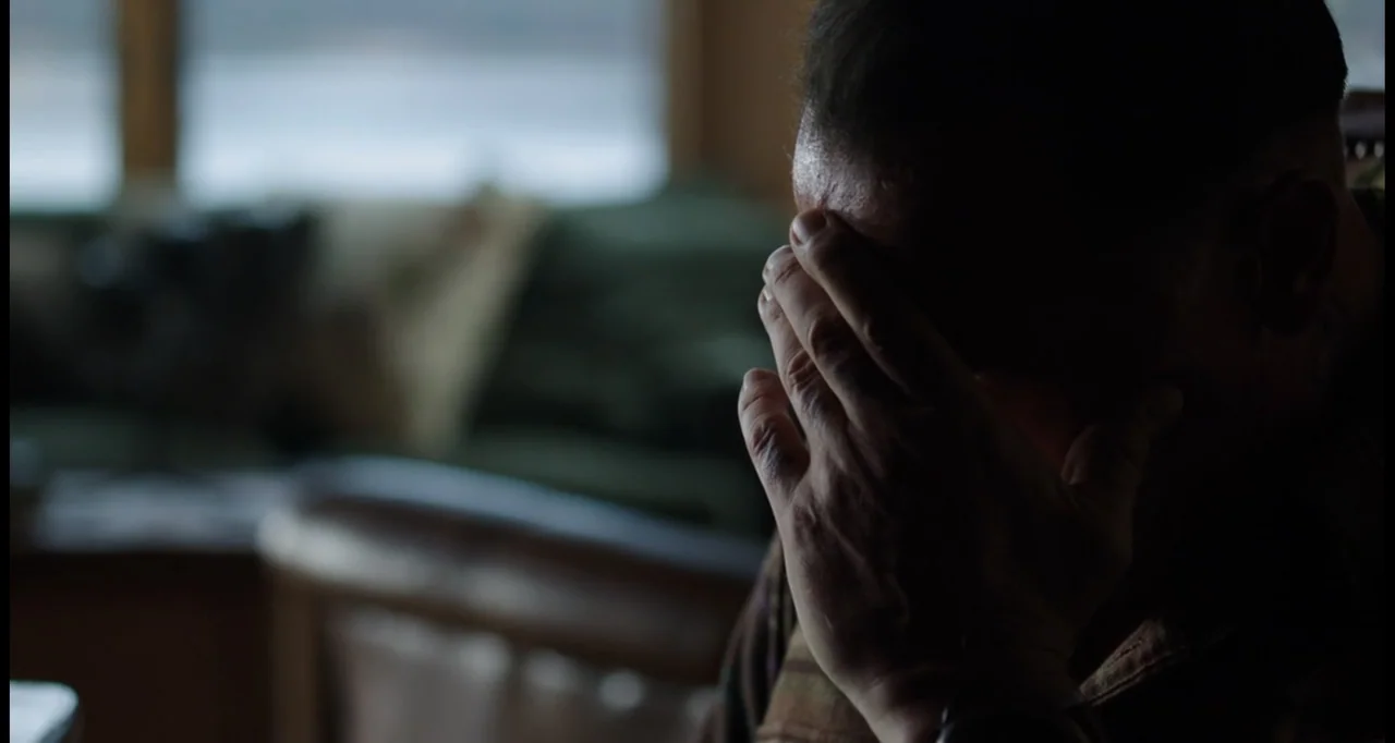 Sabotage (2014) movie still: closeup — A dimly lit room features a blurred couch visible in the background near a window. A man in a striped shirt…