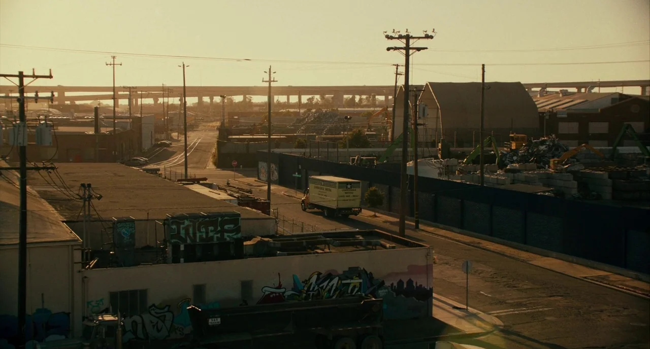 Blindspotting (2018) movie still: establishing — An industrial area at sunset features a moving truck driving past low-rise warehouses. Graffiti…