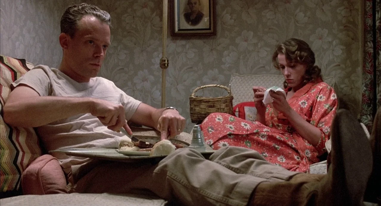 Mississippi Burning (1988) movie still: medium — A domestic space with floral wallpaper features a man and woman relaxing. The man eats a meal on a…