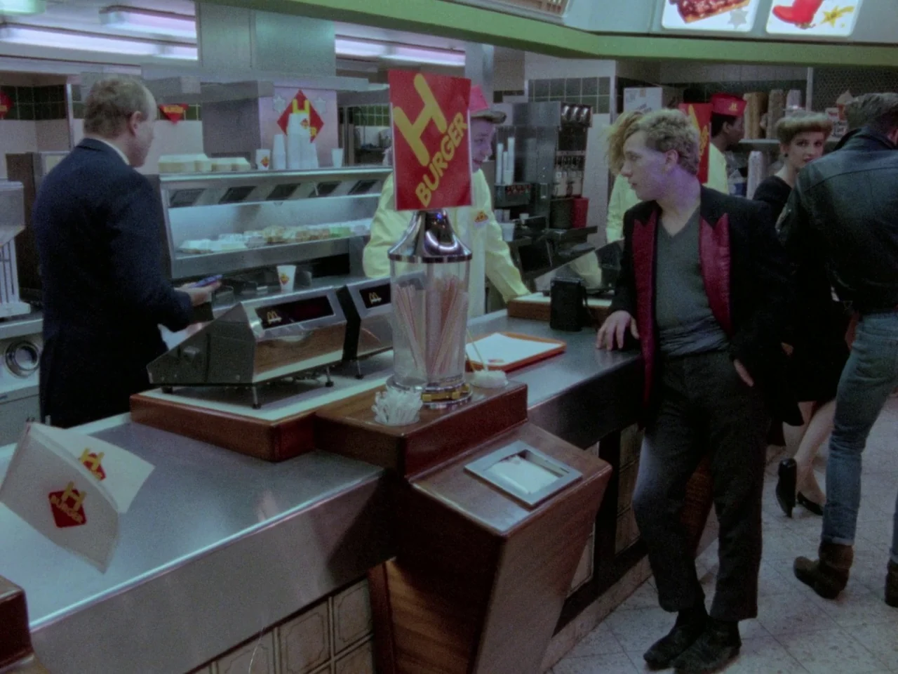 Decoder (1984) movie still: medium — An 1980s fast food restaurant interior features a stainless steel counter and menu boards. A customer leans…