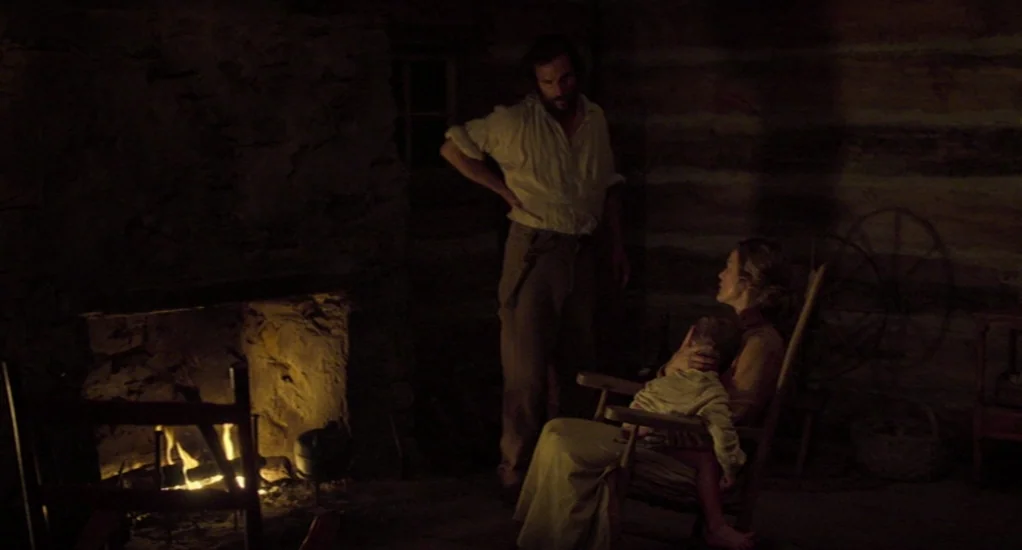 Free State of Jones (2016) movie still: medium — A dimly lit log cabin interior features rough-hewn timber walls and a large stone hearth. A man…