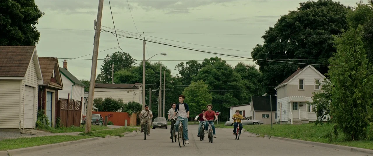 It (2017) movie still: wide — A residential road flanked by houses, trees, and power lines under an overcast sky. Five young teenagers ride bicycles…