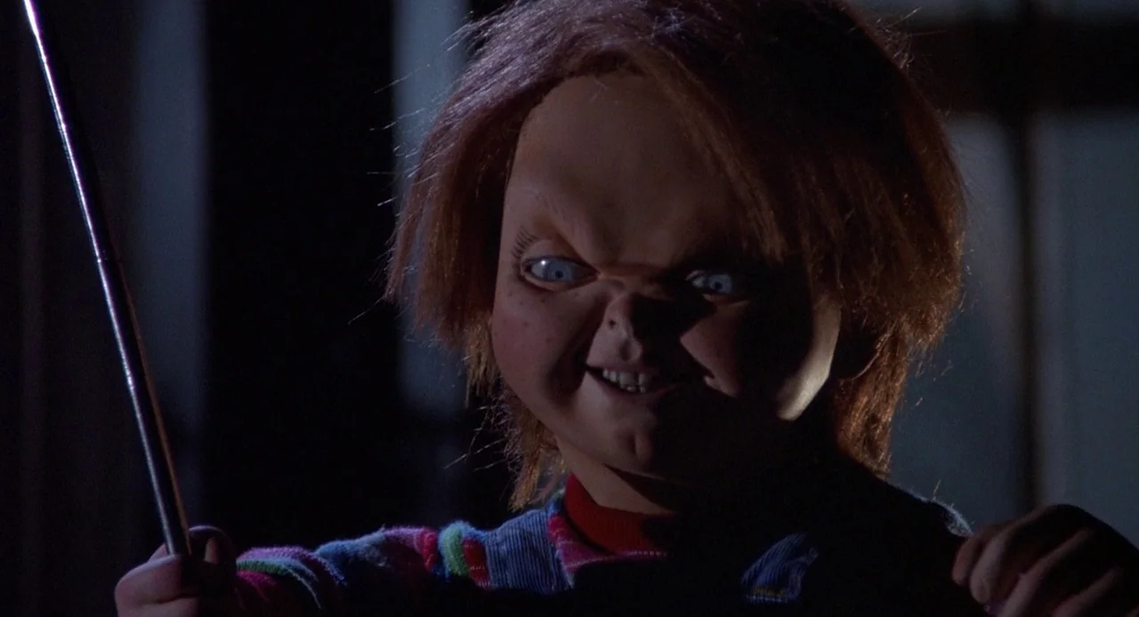 Child’s Play 3 (1991) movie still: closeup — A dark indoor space where the upper half of a doll's face is illuminated. The doll holds a slender…