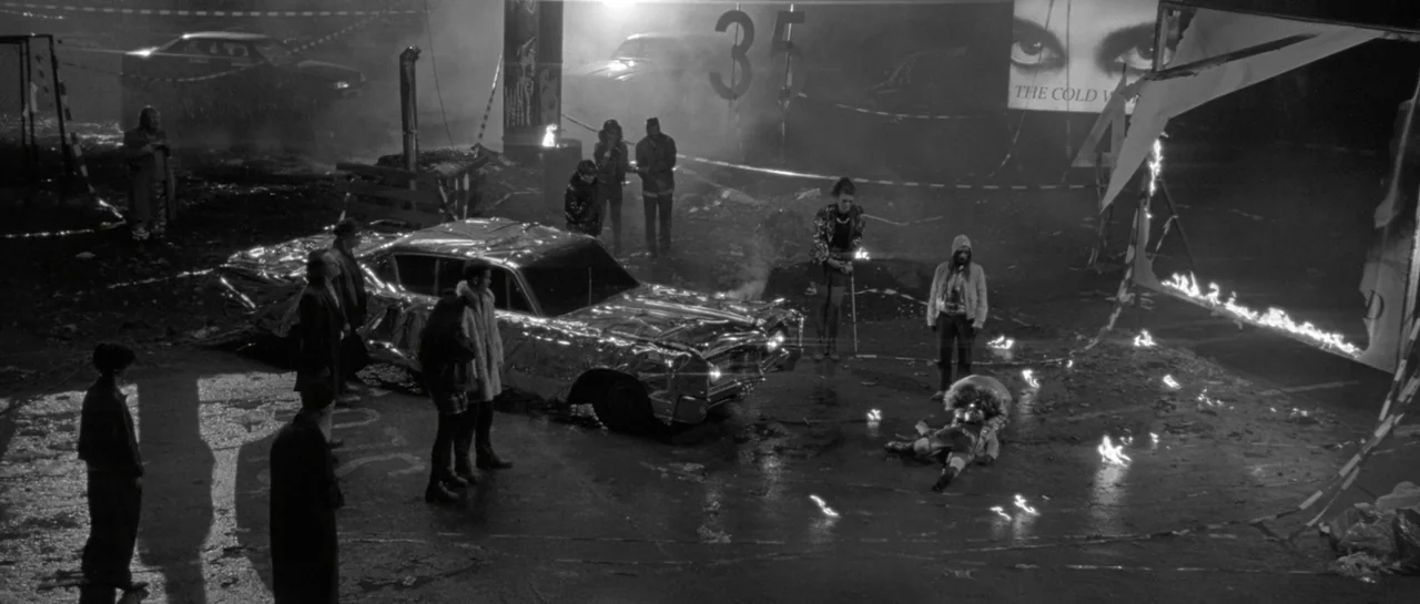 She Is Conann (2023) movie still: wide — An asphalt parking lot at night contains a car wrapped in reflective silver foil. A small group of people…