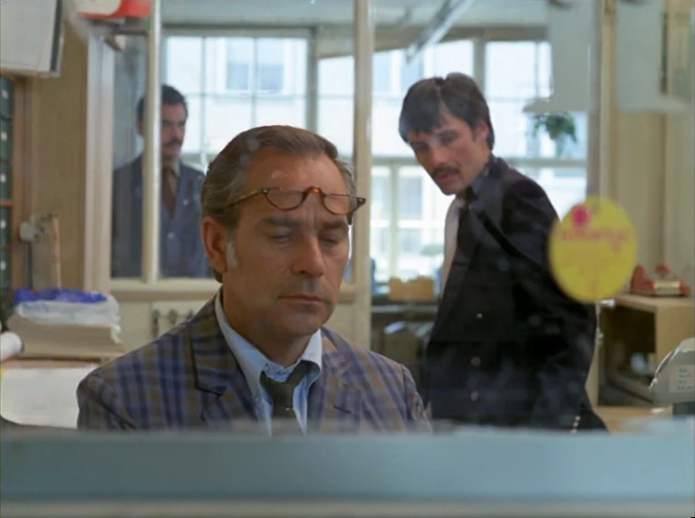 Fox and His Friends (1975) movie still: medium — A cluttered office features a desk in the foreground and windows in the back. A man sits in the…