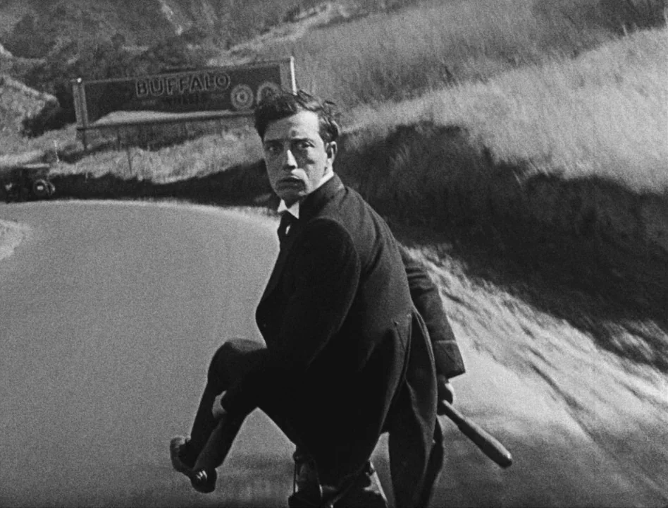 Sherlock Jr. (1924) movie still: medium — A winding road stretches through a grassy hillside with a billboard visible in distance. A man in a suit…