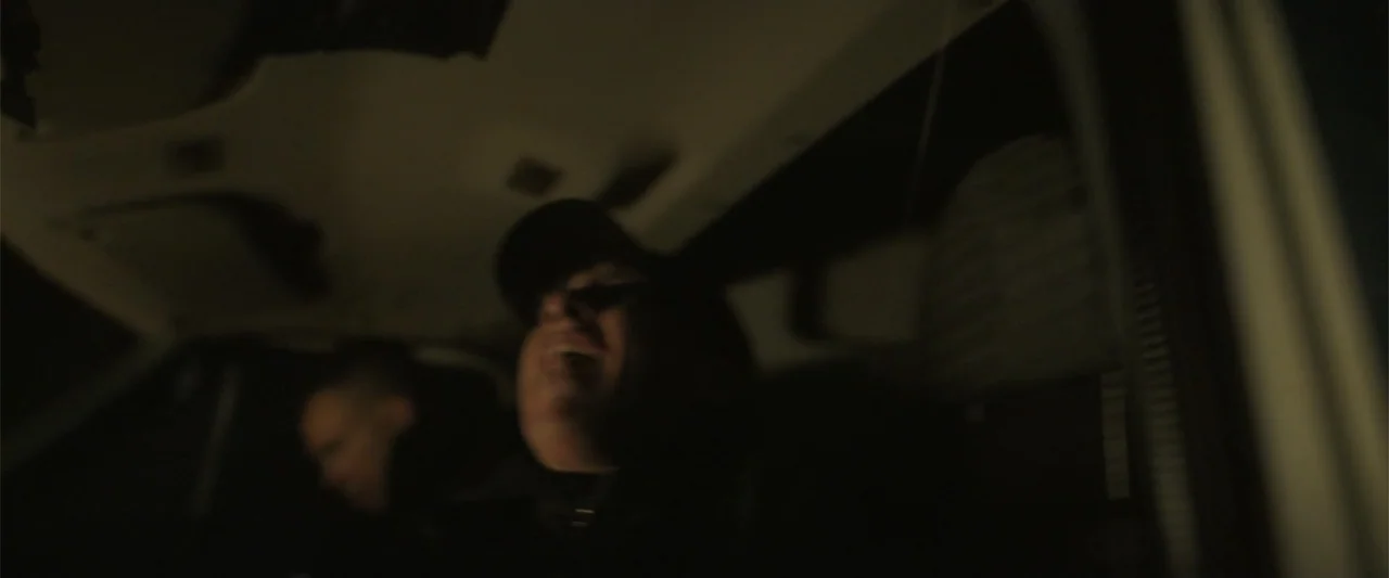 Bring Them Down (2025) movie still: medium — The interior of a vehicle at night featuring a dark cabin environment. A man in a baseball cap sits…