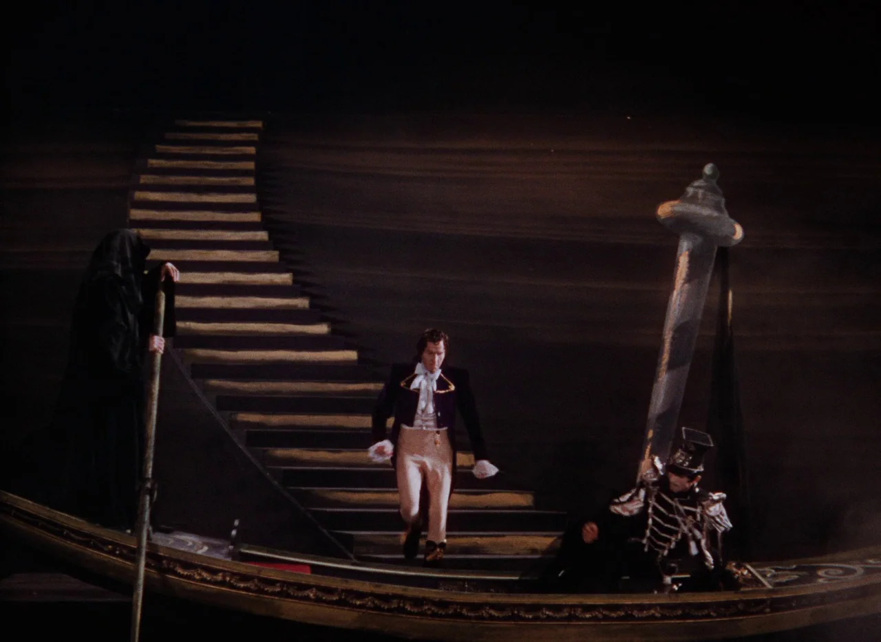 The Tales of Hoffmann (1951) movie still: wide — A surreal theatrical stage features a dark, curved staircase set against a black backdrop. A man in…