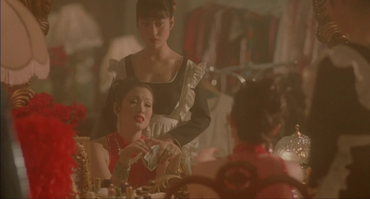 Shanghai Triad (1995) movie still: medium — A dimly lit room features a vanity table cluttered with red flowers and cosmetics. A woman sits facing a…