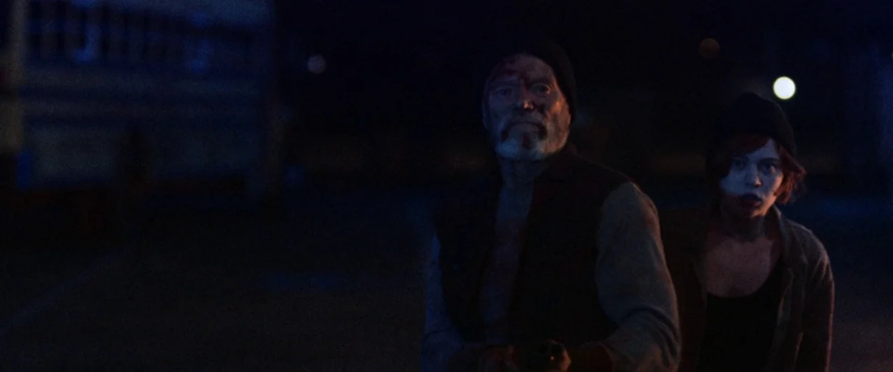 VFW movie still: medium — An outdoor location at night features a dark, expansive paved surface. An older man and a young woman stand together while…