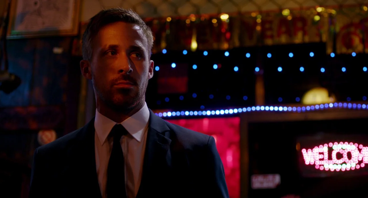 Only God Forgives (2013) movie still: medium — A dimly lit bar interior features blurred neon signage and festive decorations in background. A man…