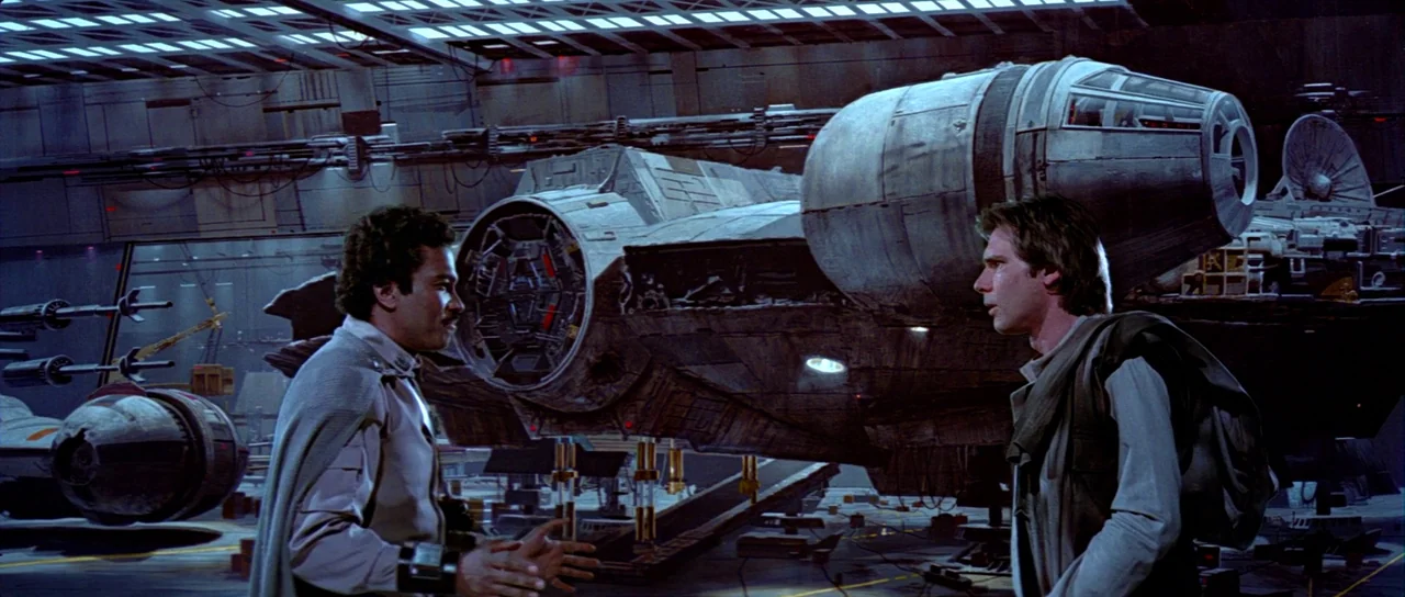 Return Of The Jedi (1983) movie still: medium — A dimly lit industrial hangar houses a large worn spacecraft with exposed machinery parts. Two men…