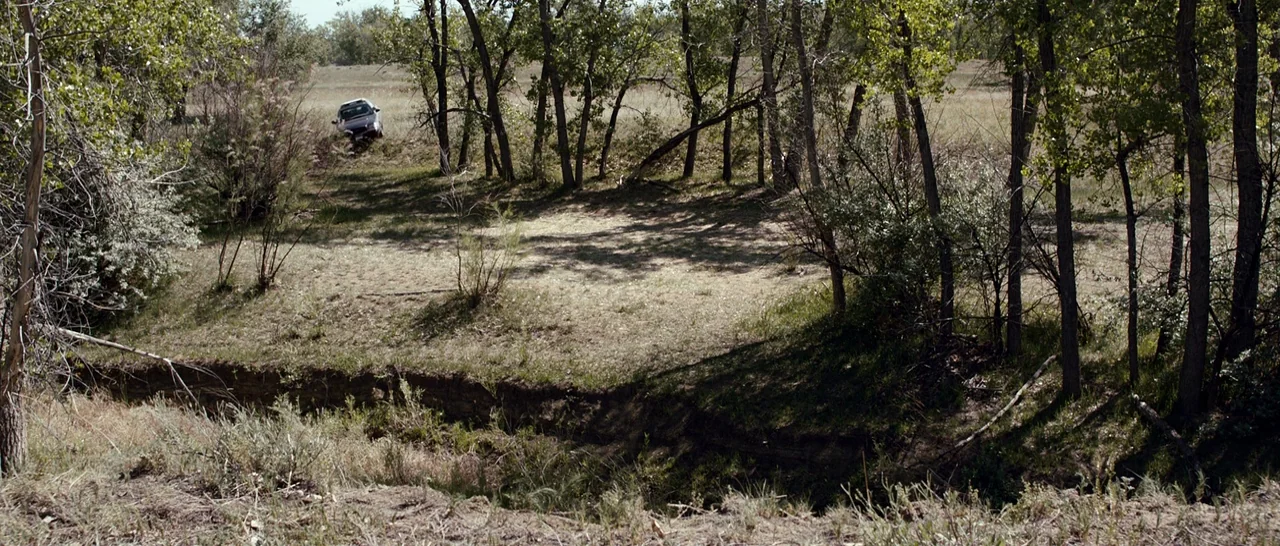 Cop Car (2015) movie still: wide — A dry, grassy meadow is bordered by a line of thin, upright deciduous trees. A single dark car is positioned on…