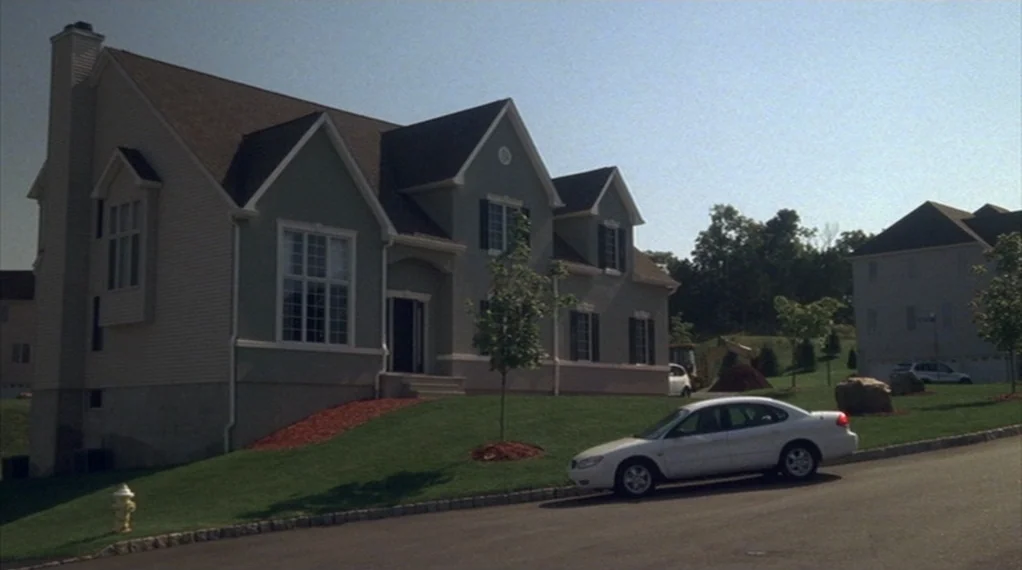 Broken Flowers movie still: establishing — A contemporary suburban house with grey siding and a lawn sits on a hill. A white sedan is parked on the…