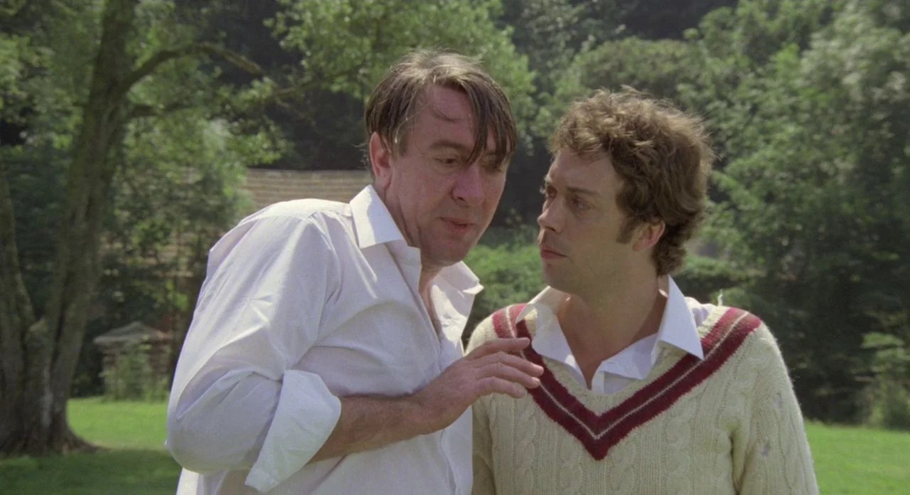 The Shout (1978) movie still: medium — Two men stand on a grassy field with mature trees in the background. The older man leans toward the younger…