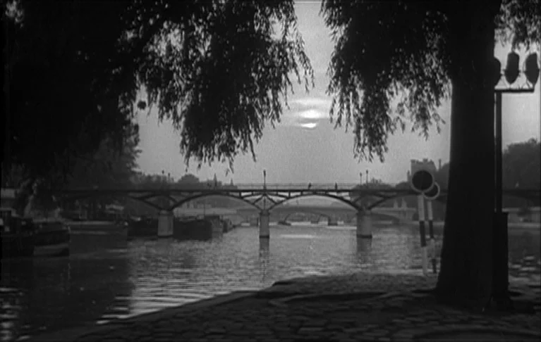 Le Signe du Lion (1962) movie still: wide — A view across a river towards a multiple-arched bridge under a twilight sky. Large tree branches frame…