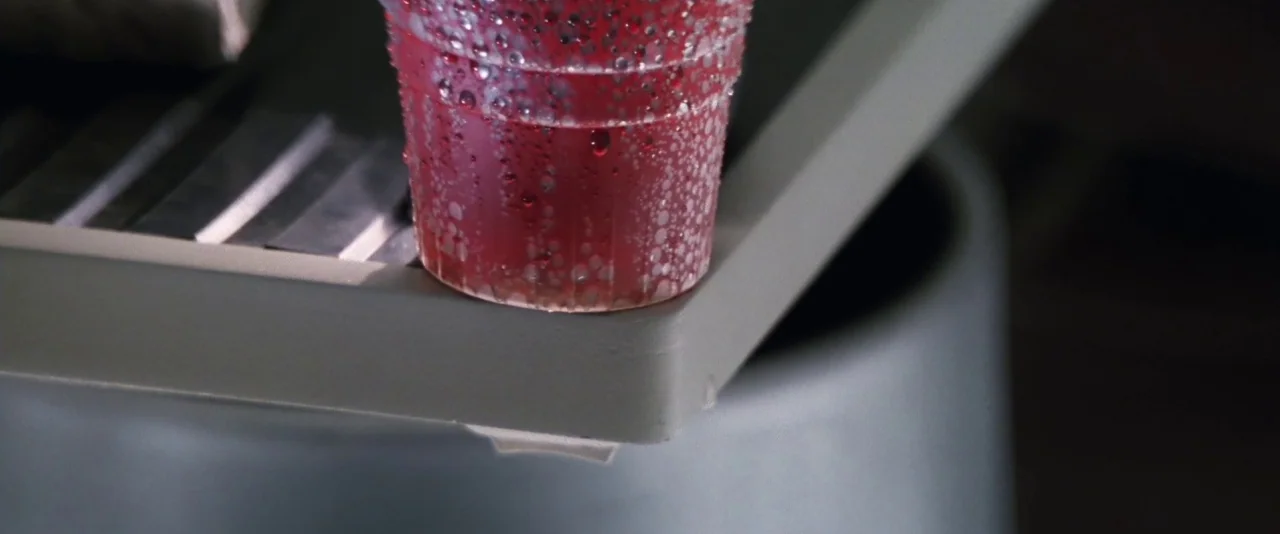 Mission: Impossible (1996) movie still: closeup — A translucent plastic cup filled with red liquid rests on a gray tray. Condensation beads cover…