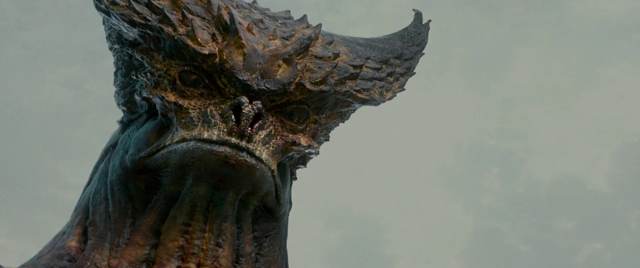 Colossal (2016) movie still: closeup — A gargantuan reptilian creature with large horns stands against an overcast sky. The subject looks directly…