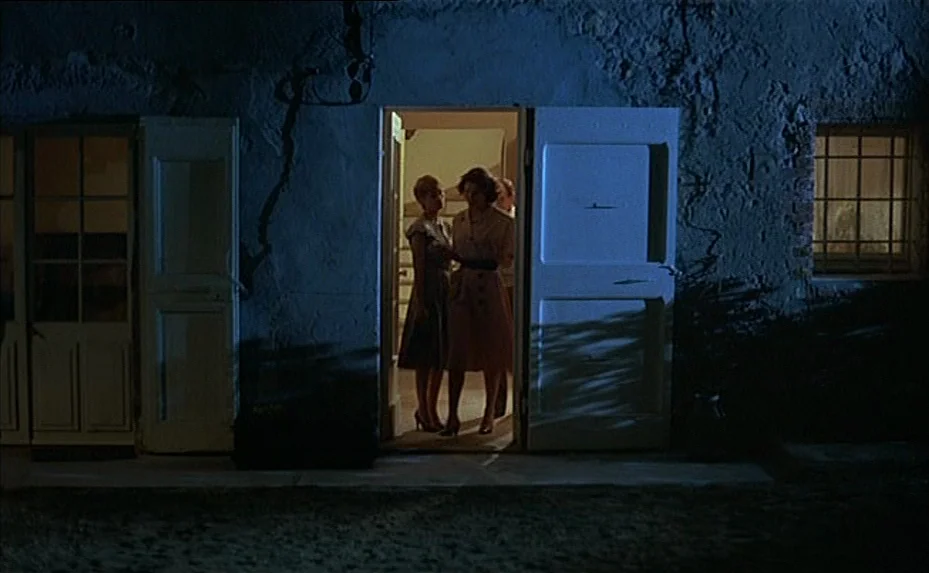 The Woman Next Door (1981) movie still: fullbody — The exterior of a stucco house at night features an open, glowing doorway. Two women stand…