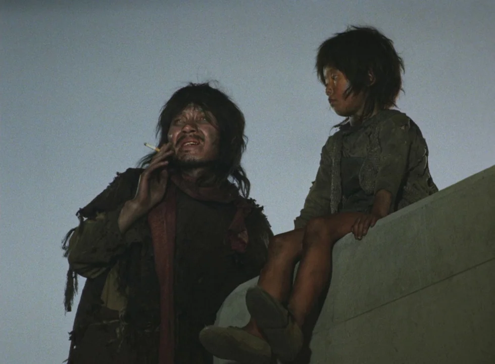 Dodes’ka-den (1970) movie still: medium — Two individuals sit atop a flat stone-like structure against a plain dusk sky. The adult male smokes a…