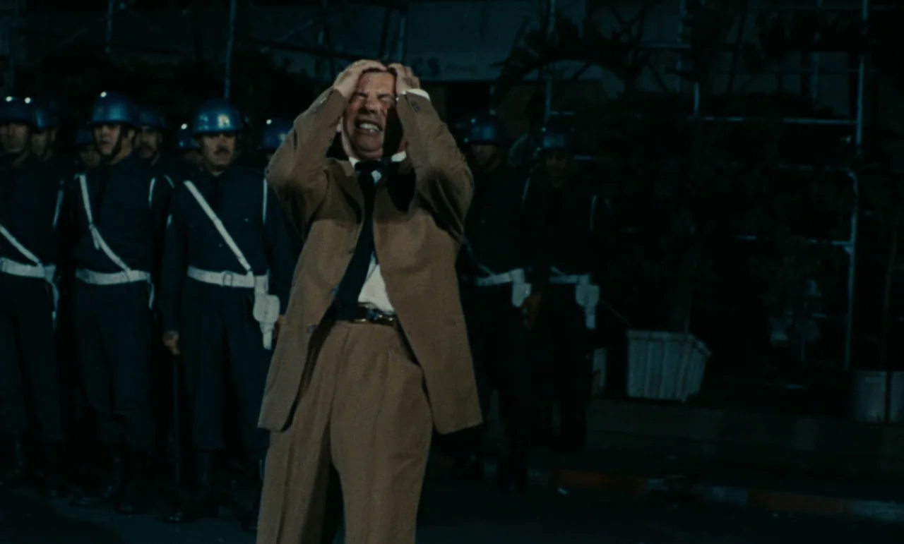 Z (1969) movie still: medium — A man stands on a dark street at night surrounded by men in uniform. The man holds his head in distress while the…